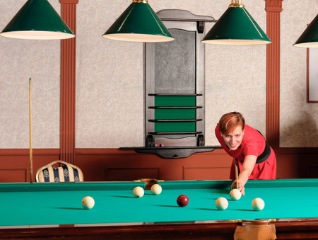 red haired woman wearing elegant red dress playing billiard in a club. woman aiming the ball with...