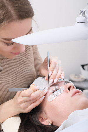 beautiful woman beautician holding tweezers with false lash to glue it to a client's eyelid....