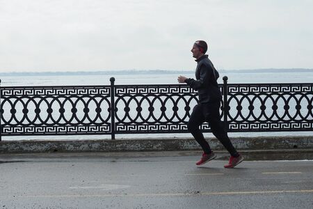 adult man is Jogging along the river on the background of a cast-iron fence on a cloudy spring morningのeditorial素材