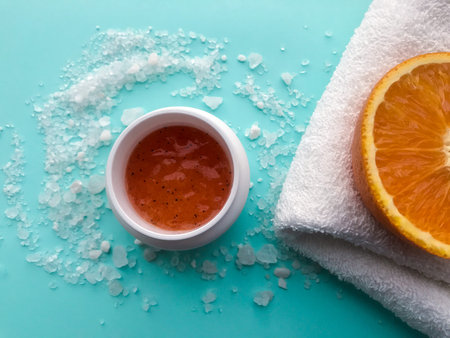 A jar of orange scrub, white towel and scattered around a large bath salt on a light blue...