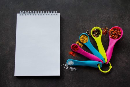 Multicolored measuring spoons with spices: chili flakes, a mix of pepper peas, curry, paprika and coarse sea salt. Next to it is a notebook with white pages and copy space. Black background.の写真素材
