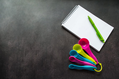 Multi colored measuring spoons, notebook with white pages and green pen for writing. Black...