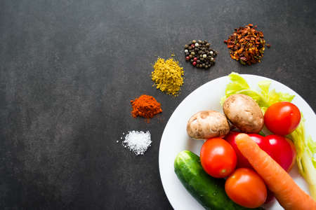 A white plate with fresh vegetables: potatoes, tomatoes, carrots and herbs, around which various spices are scattered in piles. On a black background. Copy space.の写真素材
