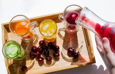 A wooden tray with colorful drinks: orange, yellow, and cucumber lemonade. Nearby are cherries. A...