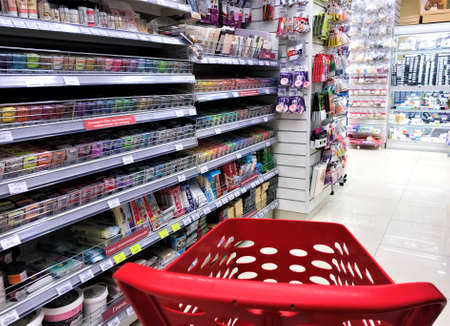 Moscow, Russia, June 2019: Close-up of empty red plastic shopping cart inside the store with stationery between narrow rows, tightly hung with small goods, opposite the shelves with polymer clay.のeditorial素材