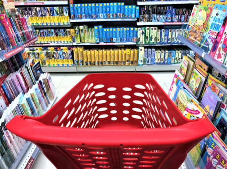 Moscow, Russia, June 2019: Close-up of a empty red plastic shopping cart inside the store between narrow rows of books and magazines, opposite the shelves with boxes of kits for assembling models.のeditorial素材
