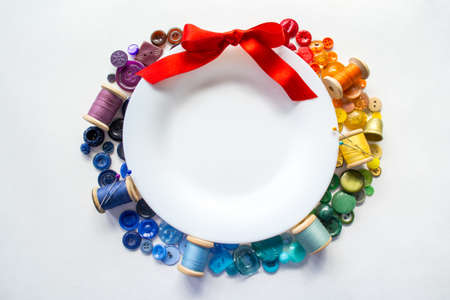 Rainbow spectrum buttons and reels with colorful threads, laid out in form of wreath with red bow around a white plate with space for text on a white background. Holiday decor concept.の写真素材