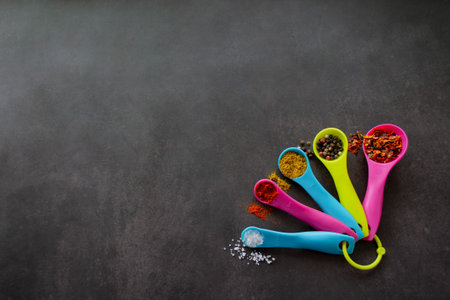 Multicolored measuring spoons with spices: chili flakes, a mix of pepper peas, curry, paprika and coarse sea salt. Black chalk board background with inscription - Herbs and spices. Copy space.の写真素材