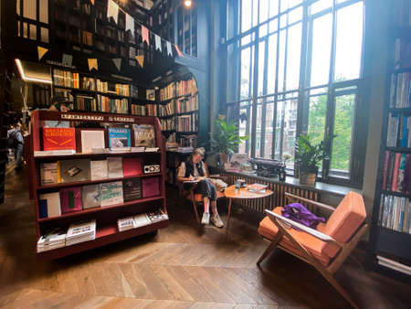 St. Petersburg, Russia, August 2021: Modern cozy bookstore. Young woman sits near window on armchair, reads, writes with small fluffy dog.のeditorial素材