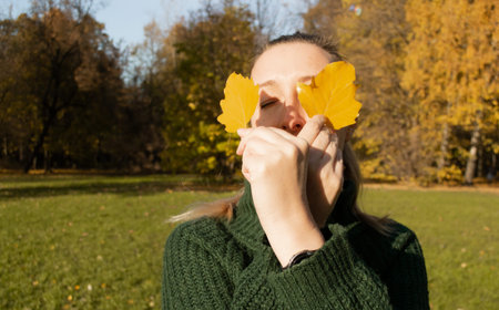 Portrait of woman with eyes closed holds two yellow leaves, hands close-up, background autumn trees.
