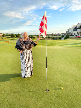 Happy laughing senior woman plays golf on golf course in evening attire. Ball is near hole with flag.の写真素材