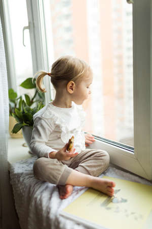 Little girl sitting with cookies on the windowsill of the house, self-isolation at homeのeditorial素材