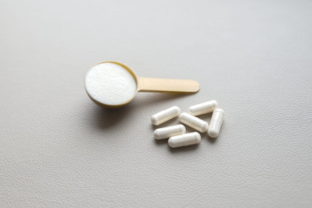 A plastic spoon of collagen powder and some pills, tablets capsules on a gray background