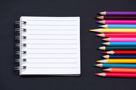 school background. Blank notepad and colored pencils on black backgroundの写真素材