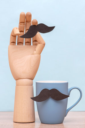Father's day concept with blue mug with mustache, wooden hand holding mustache on table. vertical photoの写真素材