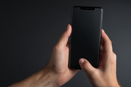 Male hand holding smartphone with blank screen on dark background, closeupの素材