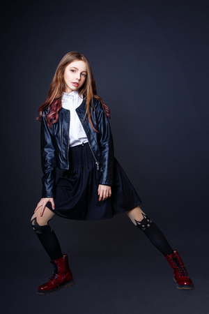 beautiful teenage girl with long hair on a black background. Studio photo. Dressed in a white blouse, black skirt, leather jacket and red shoesの写真素材