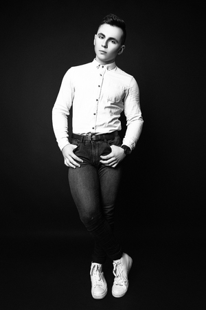 Attractive trendy young man dressed in white shirt and black jeans standing on black background. Sports guy. Studio photo. Black and whiteの写真素材