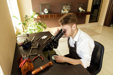 Handsome man Jeweler examines the ring through a microscope sitting at the table with tools to...