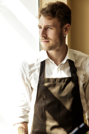 A handsome young man in an apron looks thoughtfully out the window.の写真素材