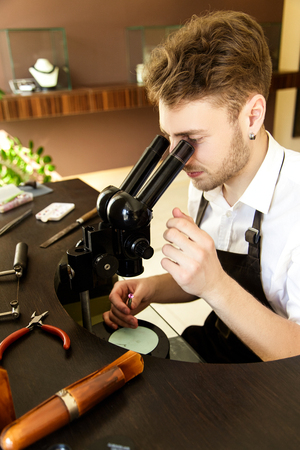 A handsome male jeweler carefully examines through a stone microscope to create a ring. Jewelry repair shop.の写真素材