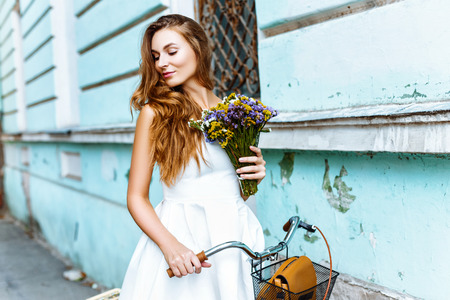 Beautiful brunette girl with long hair in white cute dress standing with yellow bicycle near blue wallの写真素材