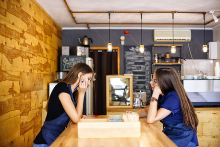 Portrait of two pensive young women sitting at a wooden table. Girls baristas in an apron are waiting for customers in a cafe. Coffee business concept. Small local business.の写真素材