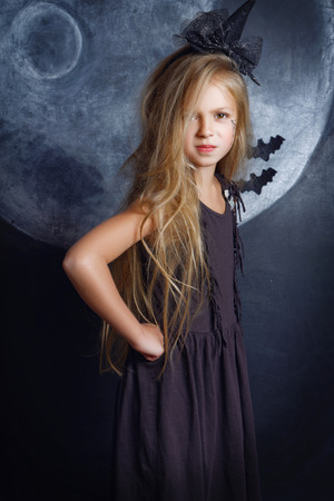 A sweet little girl with light hair in a witch's suit with a terrible look stands in the studio on the background of the moon painted with chalk on a black wall. Halloween Conceptの写真素材