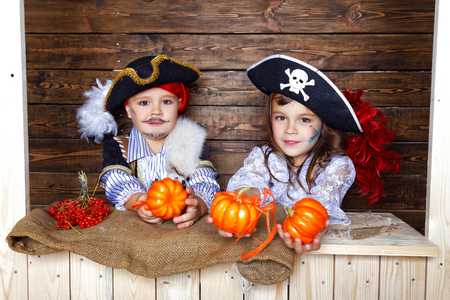 A cheerful boy and girl in pirate costumes play pumpkins near the wooden wall in the studio with Halloween decorationsの写真素材