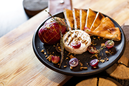 Grilled camembert with baked apple and grapes with slices of bread, served on a black plate on a wooden background in rustic style. Restaurant servingの写真素材