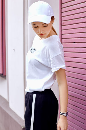 Attractive blonde dressed in a white cap, t-shirt and pants walks down the street near the pink wall...