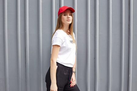 Attractive blonde wearing a red cap, t-shirt and pants stands near a gray metal wall.