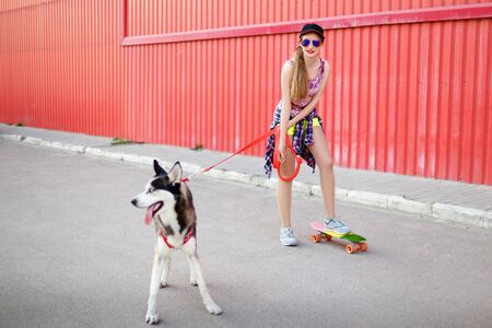 Beautiful girl in a cap and glasses skates on a skateboard with a husky dog on a background of a red wall. Summer restの写真素材