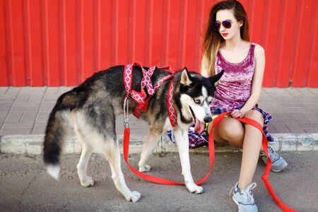 Beautiful girl in a cap and glasses skates on a skateboard with a husky dog on a background of a red wall. Summer restの写真素材