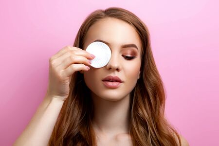 Portrait of a beautiful girl holding a sponge and cleaning makeup in the studio on a pink backgroundの写真素材