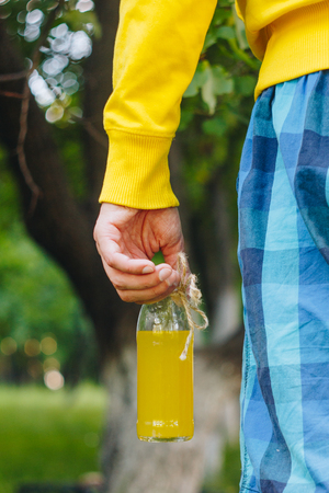 man holds in his hand bottle of homemade lemonade on the background of trees in the park and green grass. sunny day, summer. closeup healthy food fresh. cropped imageの写真素材