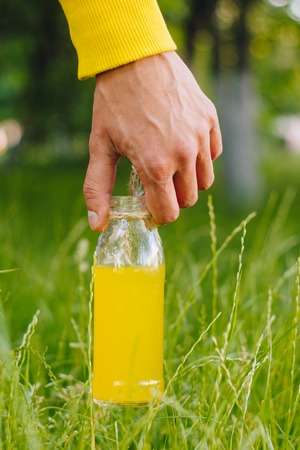 man holds in his hand bottle of homemade lemonade on the background of trees in the park and green grass. sunny day, summer. closeup healthy food fresh. cropped imageの写真素材