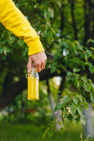 man holds in his hand bottle of homemade lemonade on the background of trees in the park and green grass. sunny day, summer. closeup healthy food fresh. cropped imageの写真素材