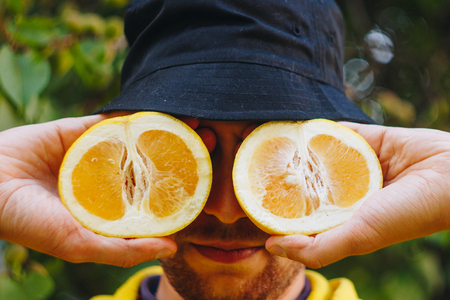 a man in a yellow hoodie and blue panama is holding two halves of a yellow citrus cut in half before his eyes. healthy eating, proper nutrition, organic food, close-up.の写真素材