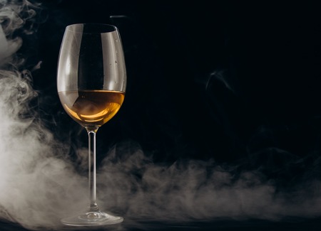 a glass of white wine on a black background. the smoke from the hookah envelops the glass. rest, holiday. alcoholic drink closeup. white fog.の写真素材