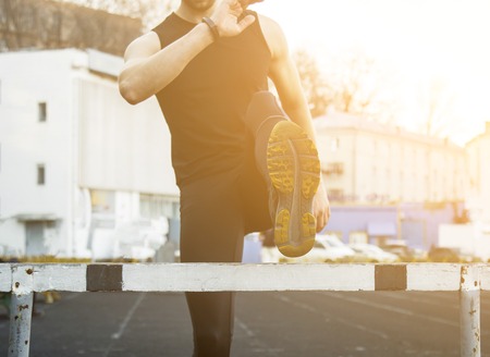 A man in black clothes is exercising outdoors with a barrier. fitness athlete on the sports field. training with hurdle. warm up stretching legs. body preparation for the summer.の写真素材