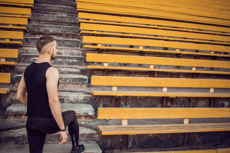 A slender young man in black sportswear stands in front of the stairs stadium stands. ready for...