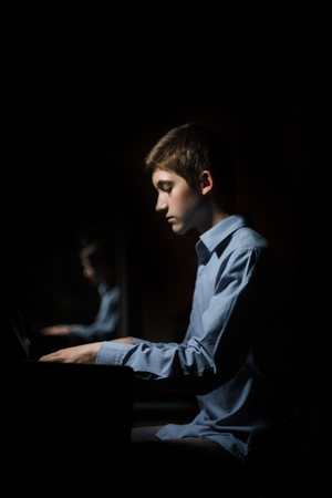 young man sitting at the piano. boy emotionally plays the keyboard instrument in the music school. student learns to play. hands pianist. black dark background. vertical.の写真素材
