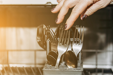 Female hand lays the dishes in an open dishwasher close-up clogged with clean, washed dishes. dry cutlery closeup. spoons forks. mugs, plates. household appliances in the kitchen.の写真素材
