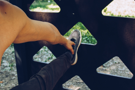 male athlete in running sports shoes standing stretches his leg on the sports field with a metal ladder. stretches hand to foot. workout first-person view.の写真素材