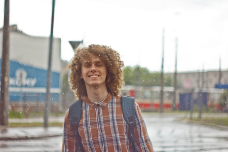 curly-haired young guy stands on the street with a satchel on his shoulder. student and young...