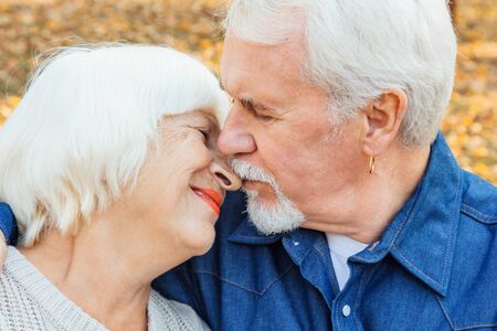 Happy elderly man and woman sitting on a bench in autumn day. Relaxed senior couple sitting on a park bench. Grandfather gently kisses grandmother on the foreheadの写真素材