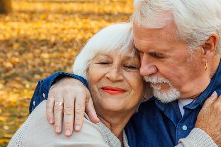 Happy elderly man and woman sitting on a bench in autumn day. Relaxed senior couple sitting on a park bench. Grandfather gently kisses grandmother on the foreheadの写真素材