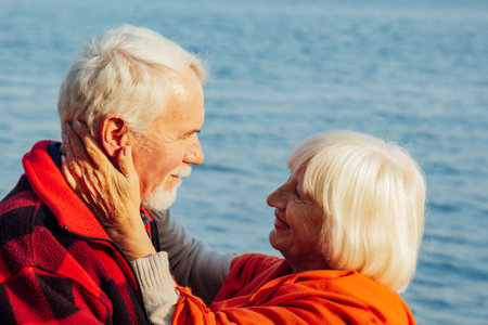 cheerful senior citizens woman and man are standing and hugging on the lake, against the background...