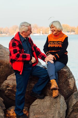 Cheerful elderly people a woman and a man are sitting on the stones and hugging on the lake, against the background of the bridgeの写真素材